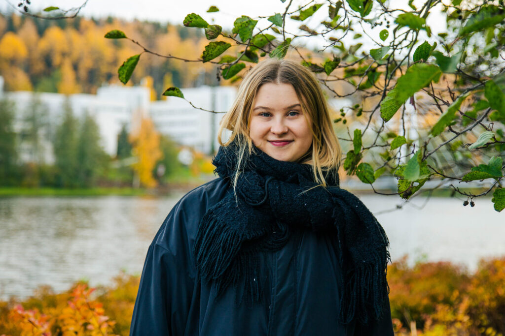 Nuori nainen katsoo kameraan, taustalla syksyinen järvimaisema.