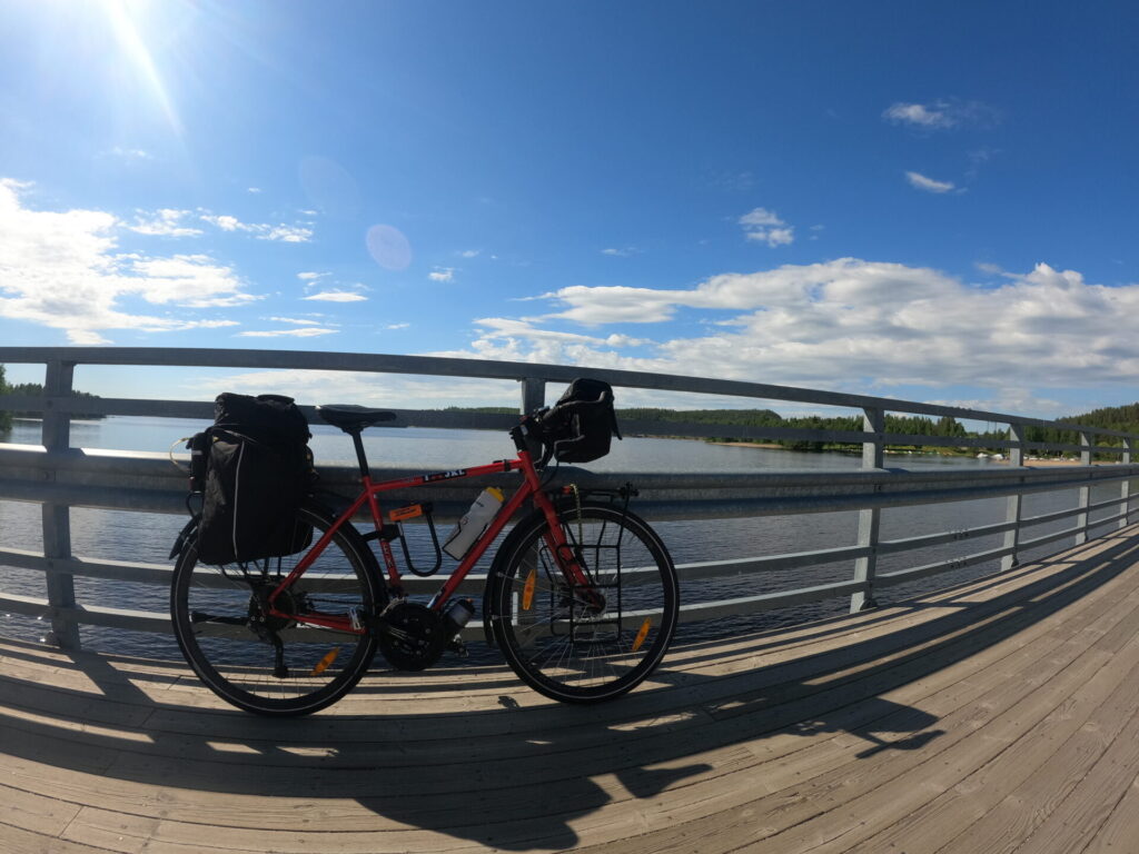 Polkupyörä nojaa sillankaidetta vasten. Taustalla järvi, sininen taivas ja pilviä.