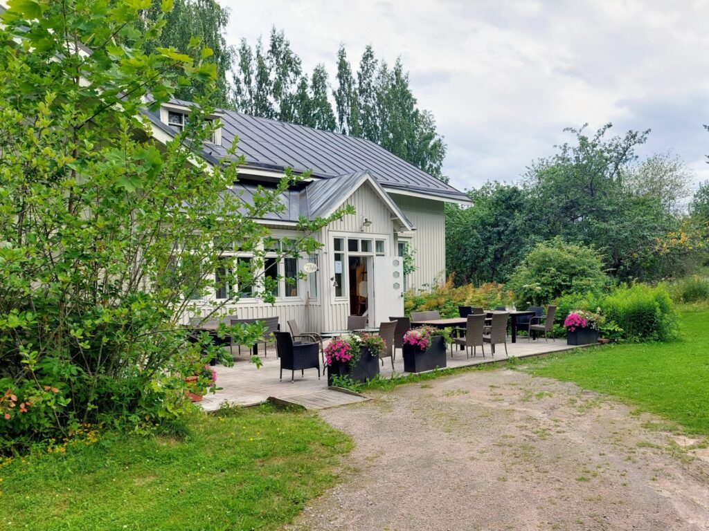 Valkoinen puutalo, ovi auki, talon edessä pöytäryhmiä.