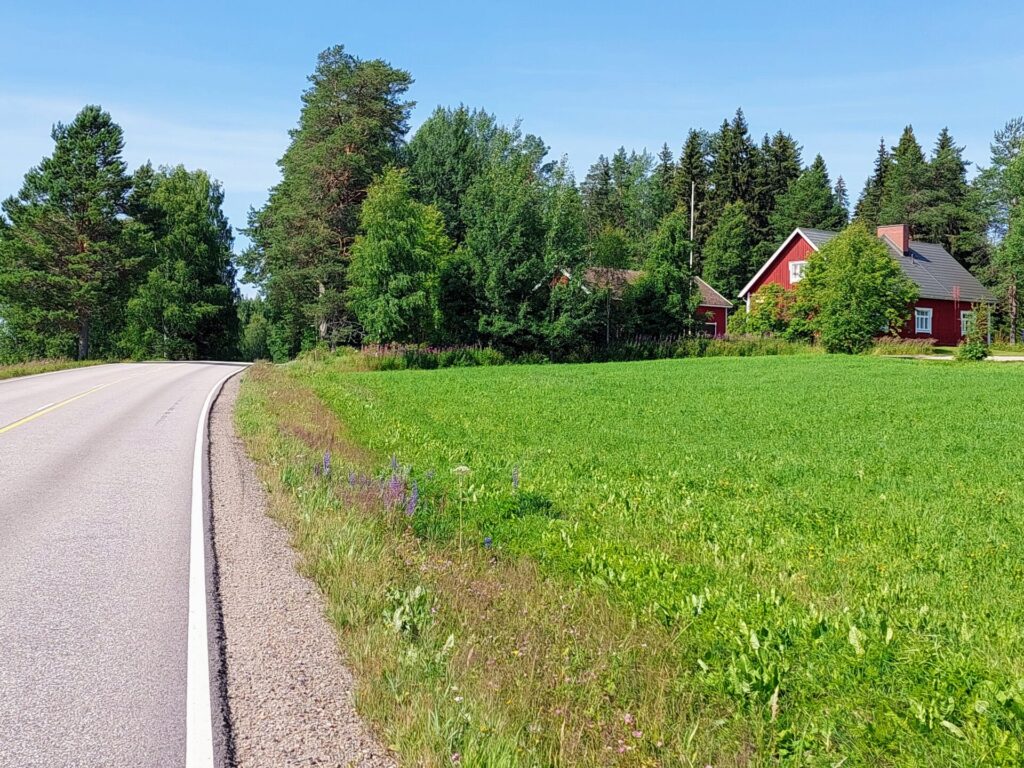 Maalaismaisema, vasemmalla autotie, oikealla pelto, takana punainen puutalon.