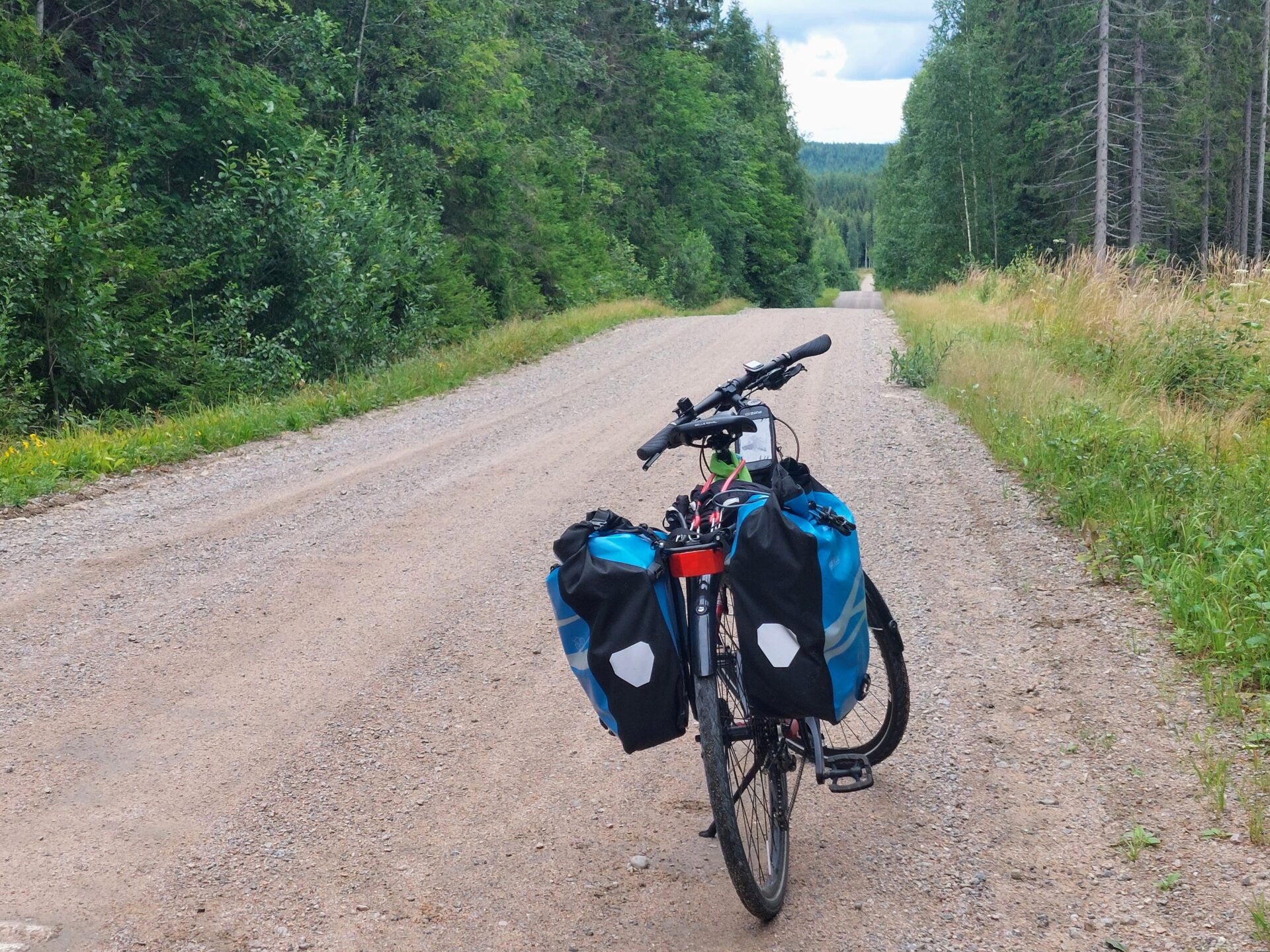 Hiekkatie, molemmin puolin metsää, tiellä pyörä, jossa kaksi sinistä pyörälaukkua
