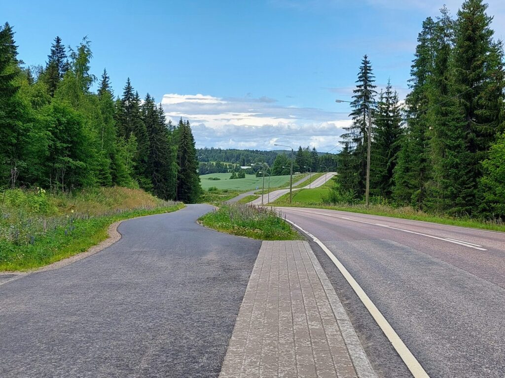 Auto- ja pyörätie rinnakkain, kumpuilevaa maalaismaisemaa, kuusimetsää