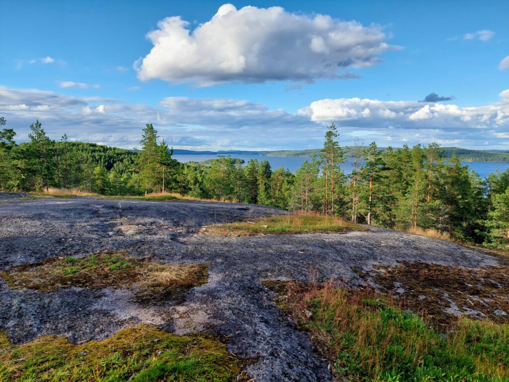 Näköalapaikka, edessä kalliota taustalla puita ja järvi, sininen taivas ja valkoisia pilviä