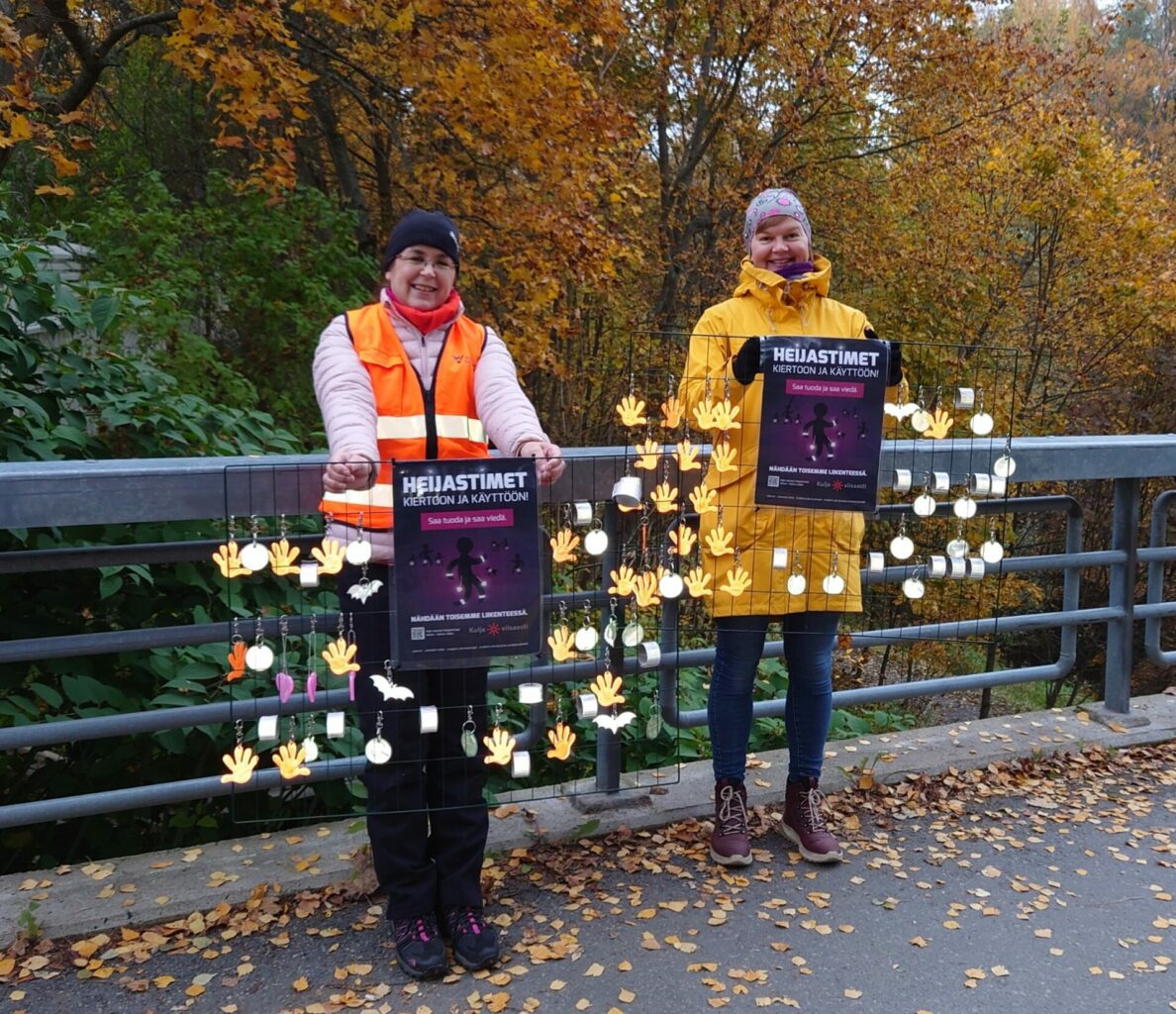 Syksyinen kuva. Kaksi naista seisoo sillalla, käsissä kehikot, joissa heijastimia. Takana keltaisia lehtipuita.